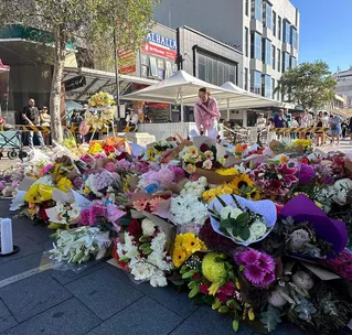 A large memorial with colorful flower bouquets on a city street, surrounded by people observing respectfully.